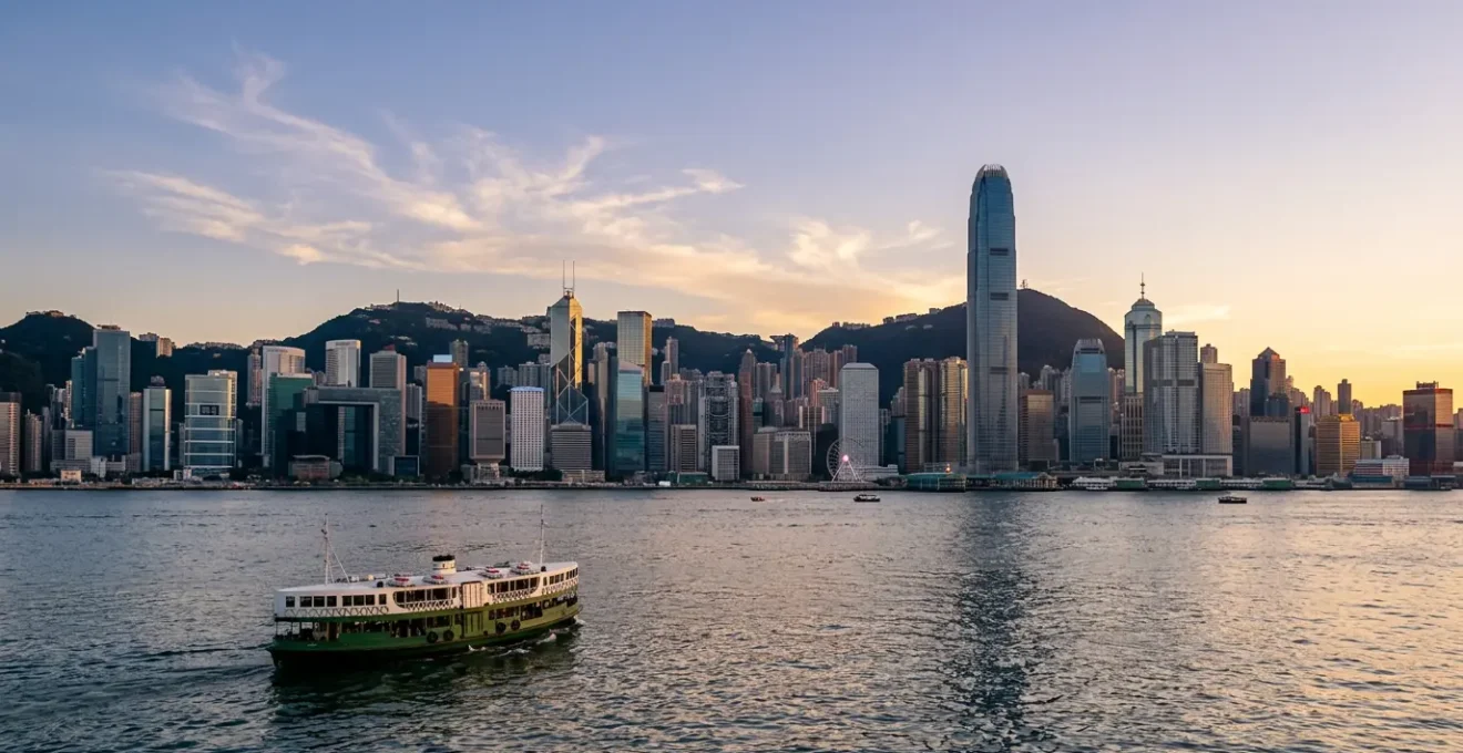 Wide panoramic view of Hong Kong's iconic skyline across Victoria Harbour during golden hour, with traditional Star Ferry in foreground