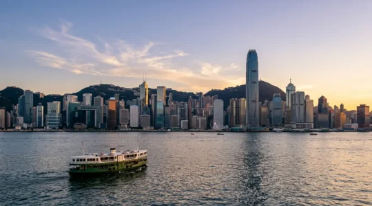Wide panoramic view of Hong Kong's iconic skyline across Victoria Harbour during golden hour, with traditional Star Ferry in foreground