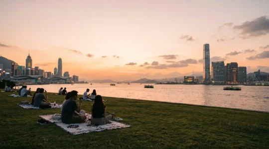 Panoramic view of people enjoying a sunset picnic on the grassy lawn at West Kowloon Art Park with Victoria Harbour and Hong Kong Island skyline in the background