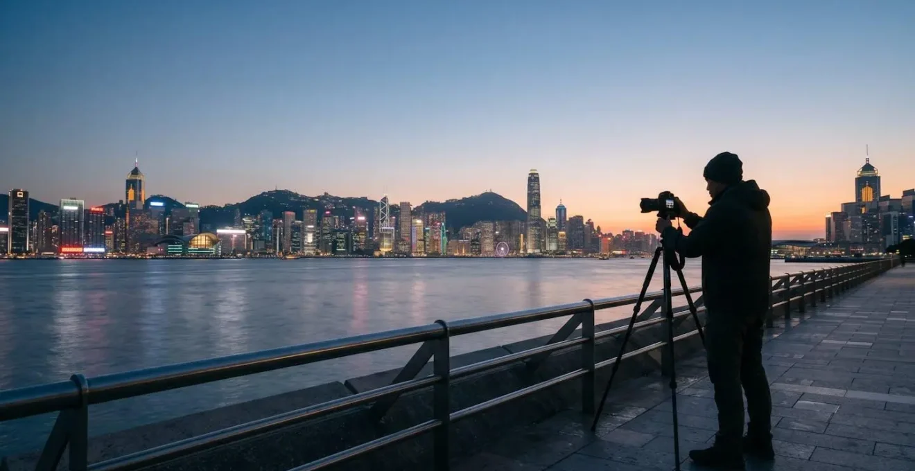 Wide environmental shot of photographer silhouette setting up camera tripod along waterfront promenade during blue hour twilight